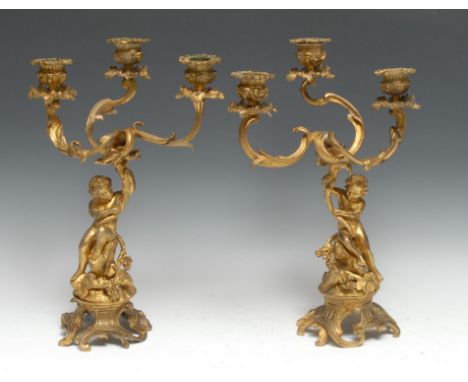 A pair of Rococo ormolu figural three-branch candelabra, cast with a pair of amorini, each seated on a rocky outcrop and hold