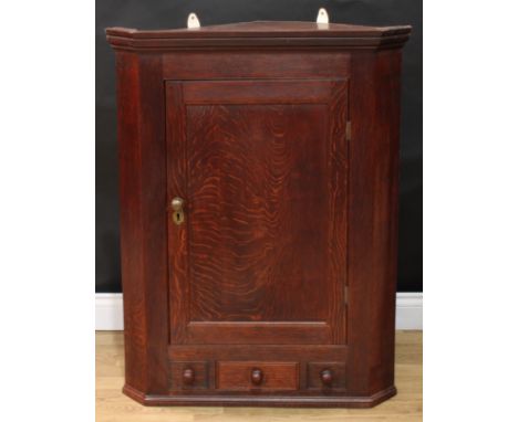 A George III oak splay-fronted wall hanging corner cupboard, moulded cornice above a rectangular panel door enclosing shaped 