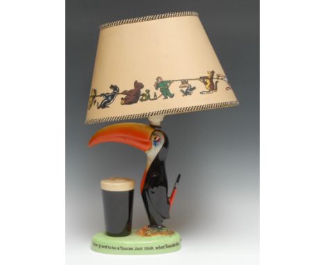 A Carlton Ware Guinness Toucan table lamp, modelled with a toucan standing before a pint of Guinness, the oval green base ins