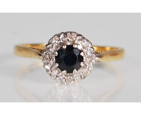 A hallmarked 18ct yellow gold ring set with a central round faceted cut sapphire with a halo of diamonds set to a decorative 