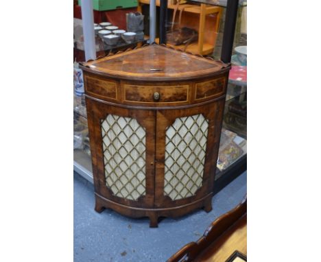 A laburnum-veneered corner cabinet with crossbanded top above bowfront drawer and two doors with brass lattice grilles, on br