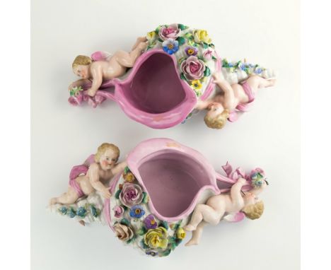 A pair of German porcelain shell wall pockets, 1920s-30s, the shells with pink interiors and ribbon, held by two winged putti