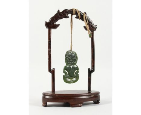 A NEW ZEALAND CARVED JADE TIKKI, suspended from a hardwood frame. Tikki: 2.75ins high.