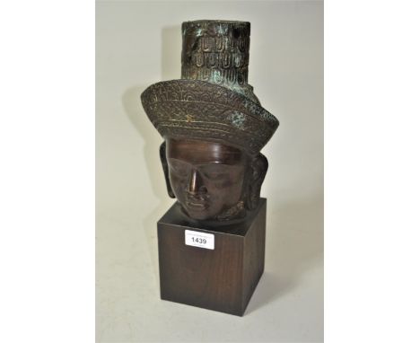 Oriental bronze head of Buddha wearing a straw hat, on a stained wooden block plinth, 14.5ins high overall 