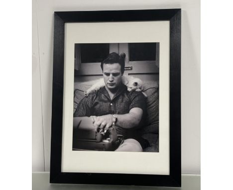 A black and white print by Murray Garr of Marlon Branco using a typewriter with a white cat around his neck, in a glazed blac