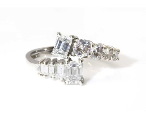 An 18ct white gold two stone emerald cut diamond crossover ring,with diamond set shoulders. Two emerald cut diamonds, each on