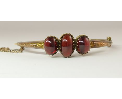 A yellow metal bangle set with three garnet cabouchons in fleur de lys galleried mounts with leaf engraved sides to the bangl