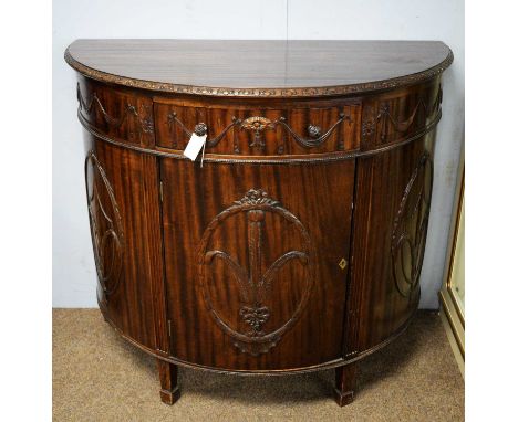 A Maple &amp; Co. demi lune mahogany side cabinet, the shaped top with carved edge above a central frieze drawer and a panell