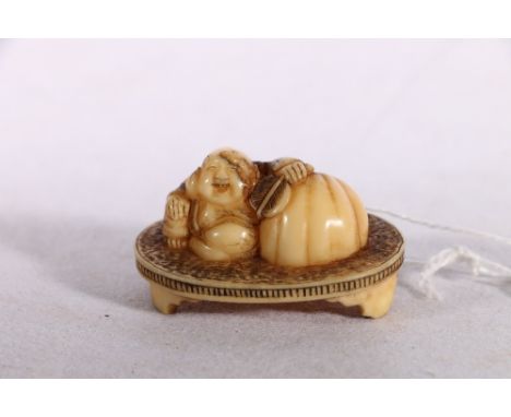 Japanese Ivory netsuke depicting Hotei holding a fan with a large bag at his side, seated on an oval table on triple supports