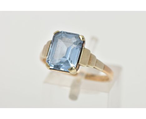 A YELLOW METAL RING, designed with an emerald cut pale blue stone assessed as synthetic spinel, step detailed shoulders leadi