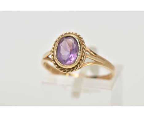 A 9CT GOLD AMETHYST RING, designed with an oval cut amethyst bezel set within a rope twist surround, trifurcated shoulders le