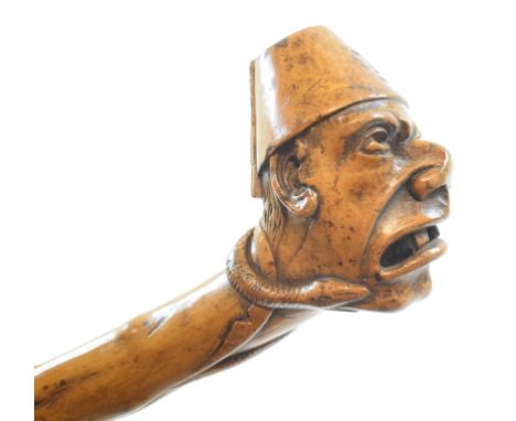A folk art walking cane, the handle in the form of a mans head wearing a fez, with his face being bitten by a snake, 96 cm 