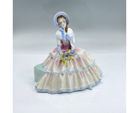 Glazed bone china figure of a seated lady in a light pink and blue dress and bonnet holding blue and yellow flowers. Part of 