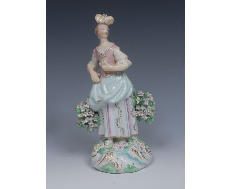A Derby Patch Mark figure, of a Flower Girl, she stands holding a posy, wearing a feathered headdress, pink bodice, blue skir