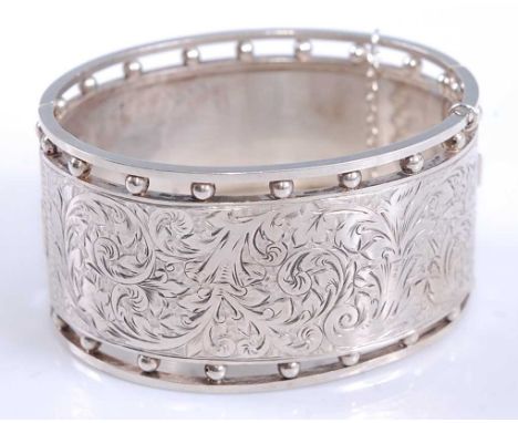 A white metal oval hinged cuff bangle, half engraved with a leaf and scrollwork design with open beaded edge, having a box cl