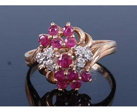 A modern 9ct gold ruby and diamond cocktail cluster ring arranged as 11 round cut rubies, dispersed with six small round cut 