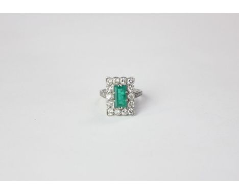 An emerald and diamond ring the step cut stone claw set in a border of fourteen round diamonds on white gold