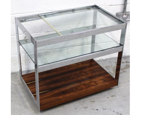 A c1970-1980 Merrow Associates glass, chrome and rosewood drinks trolley, two glass shelves above a lower rosewood shelf, wit