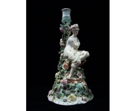 A late 19th/early 20th century German porcelain candlestick modelled as a maiden seated on a mound and amongst applied flower