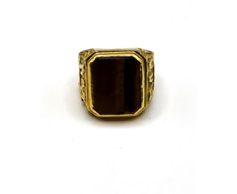 A yellow metal and tigers eye gents ring, set with a smooth tigers eye plaque, the shoulders with stylised carved decoration,