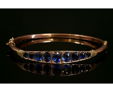 An Edwardian 9ct gold graduated synthetic sapphire and garnet-topped doublet hinged bangle, with applied leaf finials, to a h