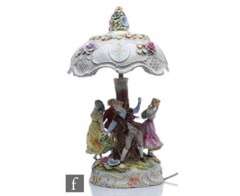 A table lamp decorated with figures dancing around a tree stump pedestal with encrusted flowers to the base, the domed shade 