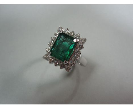 A rectangular emerald and diamond cluster ring, the emerald cut emerald four claw set within a border of eighteen round brill