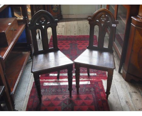 A pair of Edwardian oak hall chairs, each having pierced Gothic back, solid seat on ring turned tapering forelegs.