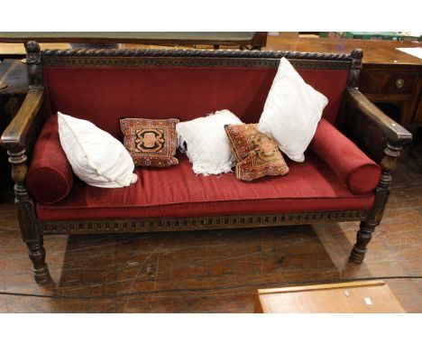 A dark oak bench with rope edge detail on back, carved figures on each end, carved legs and carved side panels. Red upholster
