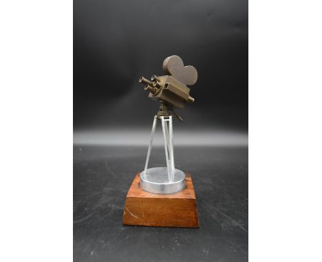 A brass and metal model of a 1930's film camera with hinged head on tripod base standing on wooden plinth. H.27 W.11cm 