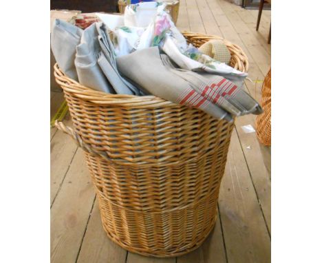 A wicker laundry basket containing a quantity of textiles including tablecloths, tray cloths, material offcuts, etc.