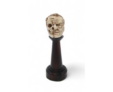 A CARVED BONE HALF SKULL/ HALF FACE with a wreath of laurel to the back of the head, upon turned wooden plinth, 11cm high Con
