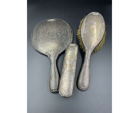 An Art Nouveau three piece silver backed dressing table set, two brushes and a mirror.