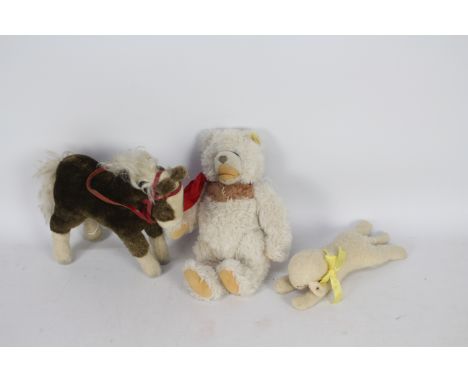 Steiff - Three Steiff plush toys. Lot includes a brown horse with red bridal and bell 25cms (H); a lying lamb with yellow rib