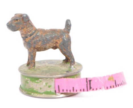 A vintage 20th century pressed metal tape measure. Having a standing dog to top, on cylindrical base with pink material tape 