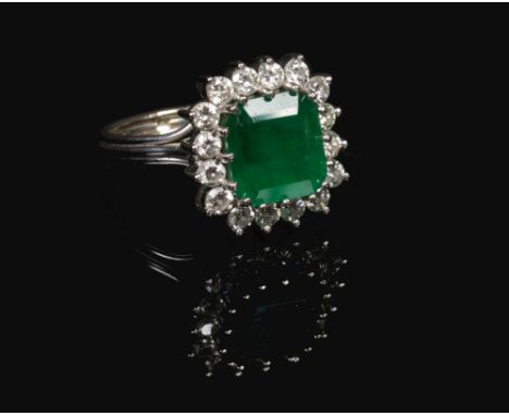 An 18 carat white gold emerald and diamond cluster ring. Set with a large square step cut emerald under a border of sixteen b