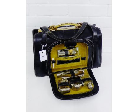 A black leather travel bag with brass frame and handles with a fall front opening to reveal a fitted interior containing a se