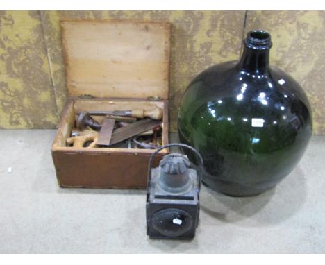 A simple wooden box containing a quantity of vintage hand tools, a railway lamp and a green glass carboy 