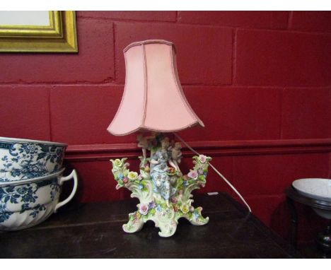 A porcelain figural table lamp depicting maiden and cherubs amongst applied flowers with pink shade    