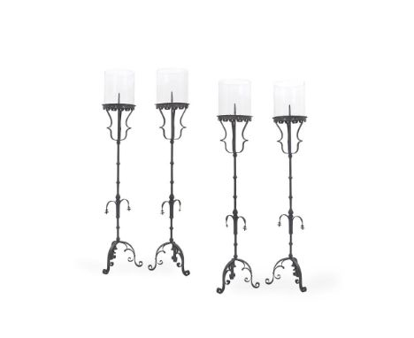 A SET OF FOUR SPANISH STYLE WROUGHT IRON CANDLESTANDS 20TH CENTURY Each with cylindrical glass storm lantern above scroll dec