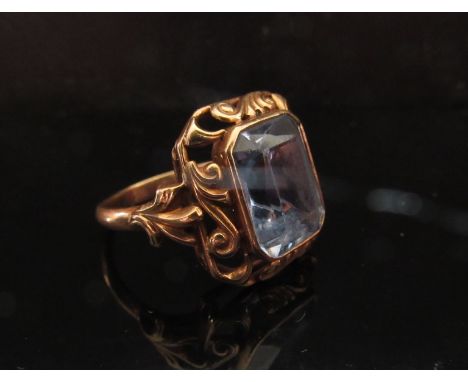 A gold ring set with an emerald cut blue stone in fancy scroll mount, marks rubbed. Size M, 5g