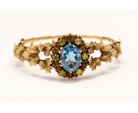 A 9ct gold hinged bangle with stone set cluster front, with leaf design decoration to the shoulders, set with an oval blue sp