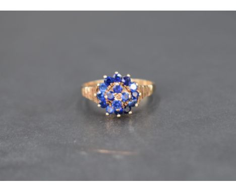 A blue gemstone set cluster ring, the central four stones with a twelve stone surround having decorative textured shoulders o
