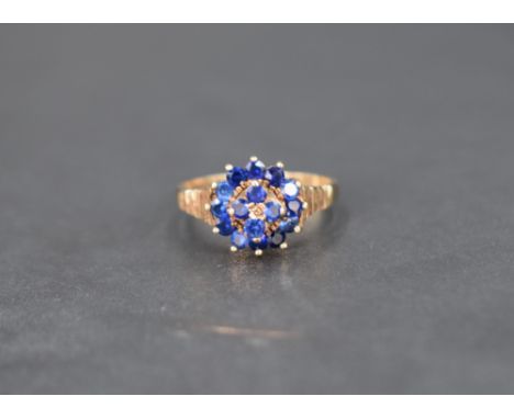 A blue gemstone set cluster ring, the central four stones with a twelve stone surround having decorative textured shoulders o