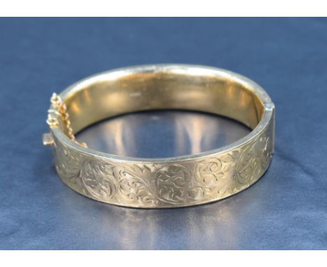 A 9ct gold hinged bangle having engraved scroll decoration, safety chain broken, inner diameter 62mm x 53mm &amp; approx 21.4