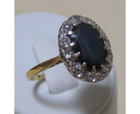 A sapphire and diamond cluster ring, the oval dark blue sapphire claw set in a border of round brilliant cut diamonds, shank 