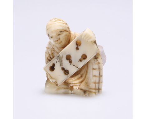 A JAPANESE CARVED IVORY NETSUKE, MEIJI PERIOD, CIRCA 1900, modelled as a seated man holding a board. Height 3.6cm