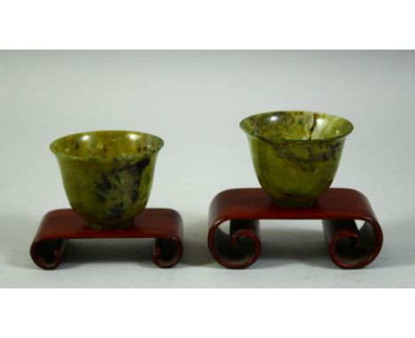 A PAIR OF CHINESE SPINACH JADE CUPS, with two wooden stands, cups 6cm diameter.