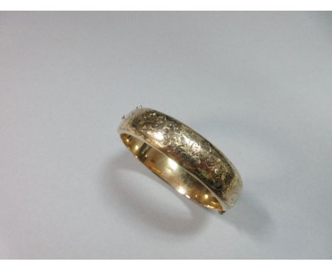 A 9ct gold engraved hinged bangle, the broad hollow D-section bangle bright cut to the front with a repeating scrolling folia