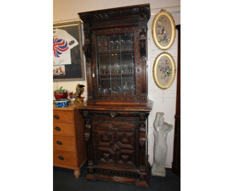 A Flemish style heavily carved oak cabinet, the upper section with lead glazed panel door enclosing a shelf, on base with fri